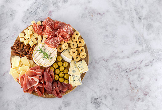 Round wooden charcuterie board with prosciutto, salami, brie, blue cheese, olives, crackers, and pecans on a marble surface